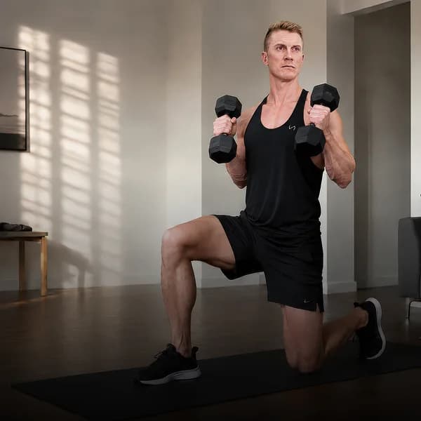 A man in a sunlit room holding a pair of dumbbells at the bottom of a lunge.